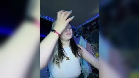 Media: A video of a young woman with light skin, long brown hair, and a white tank top, covering her face with her hand, in a dimly lit room with blue lighting and graffiti on the wall.