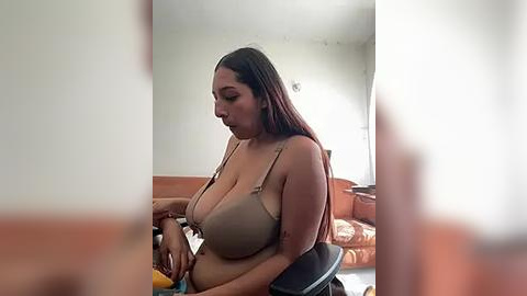 Media: Video of a plus-sized woman with long, dark hair, wearing a beige bra, sitting on a black chair in a dimly lit living room with orange sofa and white walls.