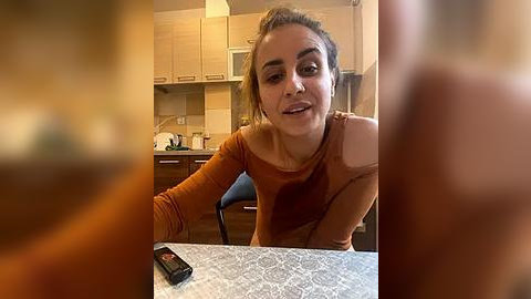 Media: A video of a young woman with light skin and brown hair in a ponytail, wearing an off-the-shoulder, rust-colored top, sitting at a kitchen table, with blurred background of wooden cabinets and beige tiles.