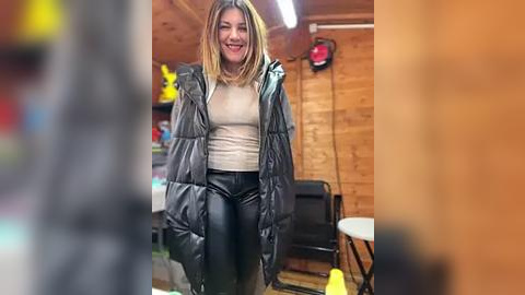 Media: Video of a smiling woman with shoulder-length blonde hair, wearing a black puffer jacket over a beige top and black leather pants, standing in a cozy, wooden-paneled garage.