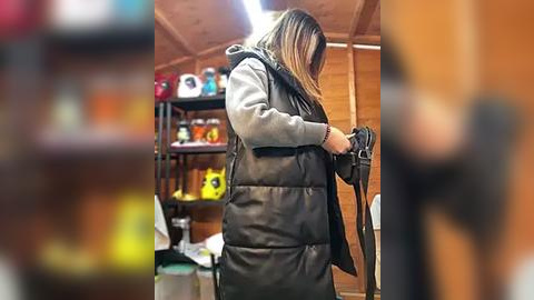 Media: A video of a woman with long hair in a black leather jacket, examining a black object in a cozy, wood-paneled room with shelves of colorful items in the background.