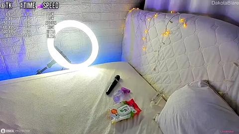 Media: Video of a cozy bedroom corner with a white quilted bed, string lights, a bright ring light, and scattered condom wrappers on a bedspread.