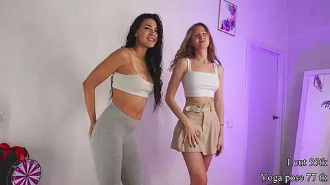 Media: Video of two young women, one with long black hair in a white crop top and grey pants, the other with long blonde hair in a white crop top and beige skirt. Background features a white wall with a door and colorful artwork.