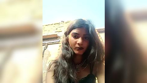Media: Video of a young woman with long, wavy black hair, fair skin, and a slender build, wearing a dark, sheer dress and multiple necklaces. She stands outdoors against a yellow building with a clear blue sky.