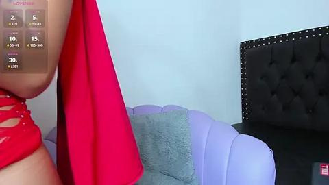 Media: Video of a woman in a red dress, holding a red object, standing beside a purple and gray tufted sofa with a black headboard.