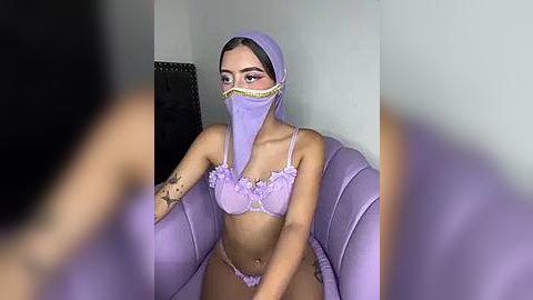 Media: A video of a young woman with light brown skin and a slim build, wearing a purple lace bra and panties, a matching veil, and sitting on a lavender couch.