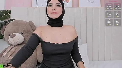 Media: A video of a fair-skinned woman with a black hijab, wearing a black off-shoulder top, seated on a bed with a plush teddy bear and white headboard in the background.