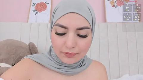 Media: A video of a young woman with fair skin, dark eyebrows, and long eyelashes, wearing a light grey hijab and a matching headscarf, with a soft, pastel pink background featuring floral wallpaper.