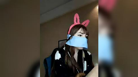 Media: Video of a woman with long brown hair, wearing a pink bunny ears headband, blue tape over her mouth, and a black hoodie, sitting in a chair indoors.