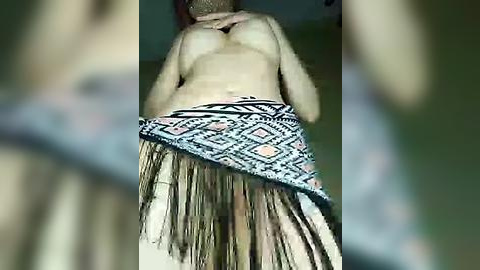 Media: Video of a shirtless man, light-skinned, with a tribal-style, fringed, multicolored sarong, partially covering his lower back, standing indoors.