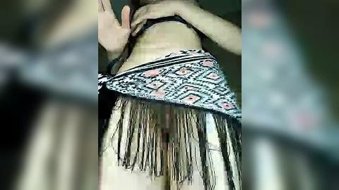 Media: Video of a woman with fair skin, wearing a black bra and a fringed, patterned skirt, partially covering her lower abdomen. The background is blurred, focusing on the garment details.