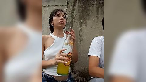 Media: A video shows a young woman with short brown hair, wearing a white tank top, holding a bottle of orange juice against a concrete wall.