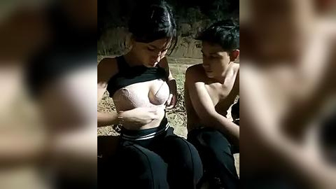 Media: A video shows a young woman with dark hair, wearing a black crop top, exposing her breast, sitting next to a shirtless young man with short dark hair. They are outdoors, dim lighting.