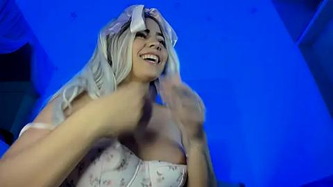 Media: Video of a smiling, light-skinned woman with long, platinum blonde hair, wearing a white floral dress, set against a blue background. She appears joyful and animated.