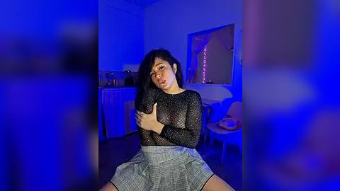 Media: Video of a young woman with long black hair, wearing a sheer black mesh top and a grey skirt, sitting in a dimly lit room with blue lighting, her hands covering her breasts.