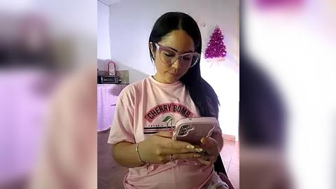 Media: Video of a young Asian woman with long black hair and glasses, wearing a pink \"Sorority Row\" t-shirt, holding a pink smartphone, in a softly lit room with a pink tree decoration on the wall.