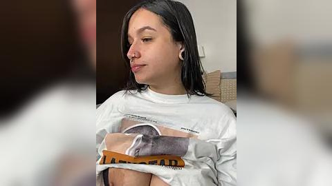 Media: Video of a young Latina woman with straight black hair, wearing a white t-shirt, holding a brown blanket over her chest, sitting on a beige couch in a dimly lit room.