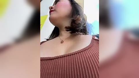 Media: Video of a young Asian woman with shoulder-length dark hair, wearing a ribbed, off-shoulder, rust-colored top, and a delicate gold necklace, captured from a low angle emphasizing her cleavage and neck. The background is blurred with yellow and blue hues.