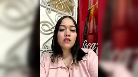 Media: Video of a young Asian woman with straight black hair, fair skin, and medium-sized breasts, wearing a light pink cardigan, seated indoors with a blurred background featuring ornate metalwork and a red sign.