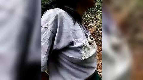 Media: Video of a person with long black hair, wearing a gray sweatshirt with a white pocket, standing outdoors against a blurred background of greenery and a brown patch.