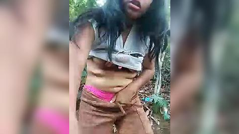 Media: Video of a woman with long black hair, wearing a torn, gray crop top exposing her breasts, pink panties, and brown shorts, standing in a forest with blurred trees and greenery.