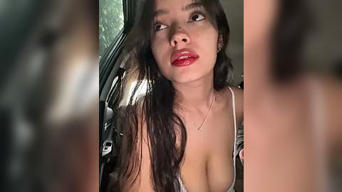 Media: Video of a young woman with long dark hair, wearing a low-cut top, sitting in a car. Her lips are red and she looks surprised.