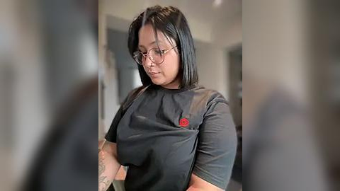 Media: Video of a young woman with medium-dark skin, straight black hair, and glasses, wearing a black T-shirt with a red button. She stands indoors, slightly blurry, in a modern room with white walls and a doorway.