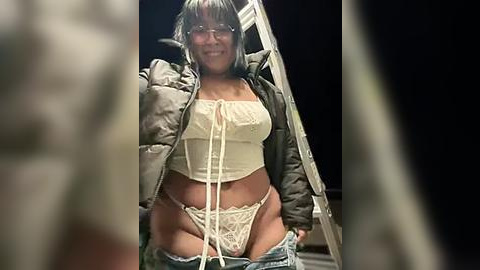 Media: Video of a smiling, middle-aged woman with glasses, wearing a beige corset top and white lace panties, unbuttoned jeans, standing next to a ladder in a dimly lit room.