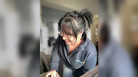 Media: A video of a young woman with black hair in a ponytail, wearing glasses and a dark sweatshirt, smiling and leaning over a wooden table in a bright, modern kitchen.