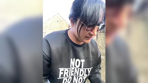 Media: A video of a young woman with dark hair and glasses, wearing a black \"NOT FRIENDLY DON'T TOUCH\" sweatshirt, standing outside in a blurry, rural setting with a beige building in the background.