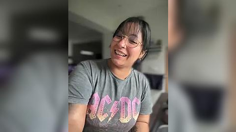 Media: Video of a smiling, dark-haired Asian woman with glasses, wearing a gray T-shirt with \"MELLODY\" in pink. Background shows blurred, indistinct people.