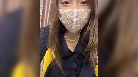 Media: Video of an Asian woman with long, straight brown hair, wearing a blue sweater with yellow sleeves, and a white lace mask, indoors, blurred background.