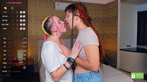 Media: Video of two women with vibrant hair, one in a white crop top, the other in a light blue top, kissing passionately in a bedroom with gold-patterned wallpaper and a bed.