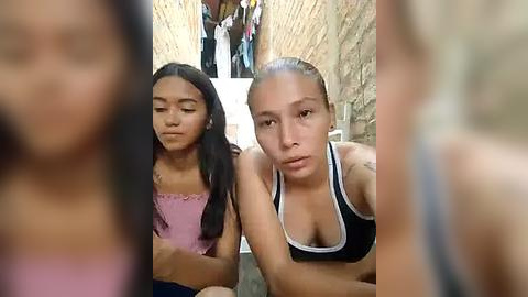 Media: Video of two young women, one with long black hair in a pink dress, the other with blonde hair in a black tank top, seated in a narrow, stone-walled alley.
