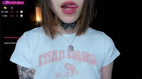 Media: Video of a young woman with pale skin, straight brown hair, and a black choker with a butterfly tattoo. She wears a white \"KISS\" T-shirt and has her tongue out, against a dark background.