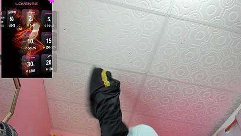 Media: Video of a person in a pink room with a white textured ceiling. The person is wearing a black jacket and a light blue shirt, holding a black bag.