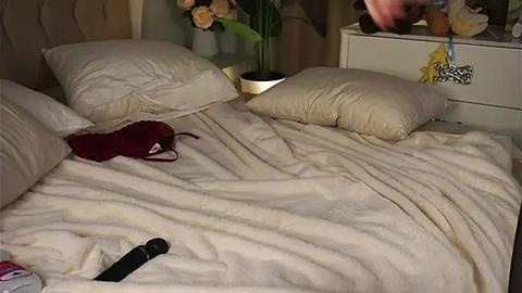 Media: Video of a dimly-lit bedroom with a beige bedspread, a red bra, and a black vibrator on the bed. The background includes a white nightstand with a potted plant and a lamp.