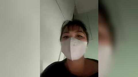 Media: Video of a woman with medium skin tone and dark hair, wearing a white surgical mask, black shirt, and standing in a dimly lit hallway with white walls.