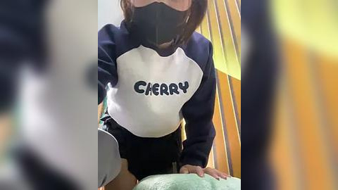 Media: A video shows a person in a white and navy blue long-sleeve shirt with \"CHERRY\" printed on the chest, wearing a black mask, and leaning over a table. The background features wooden and yellow structures.