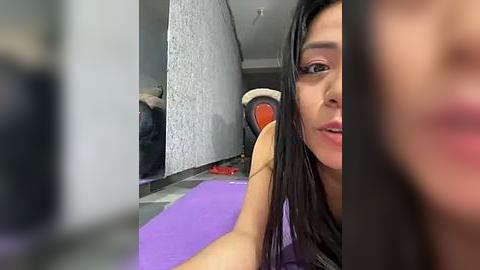 Media: Video of a young Latina woman with long black hair, fair skin, and light makeup, lying on a purple yoga mat in a gym with a white wall and orange exercise equipment in the background.