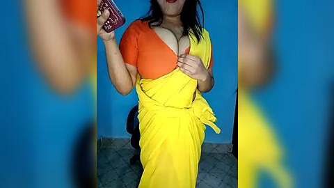 Media: A video of a woman with medium skin tone and dark hair, wearing a bright yellow sari with orange and yellow blouse, holding a red phone. Background: blue wall, tiled floor.