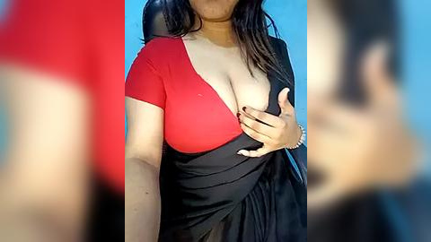 Media: Video of a woman with medium skin tone, wearing a red and black sari, holding her breast. Background is blurred, showcasing a bright blue sky.