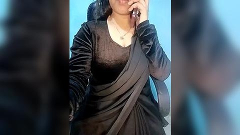 Media: Video of a South Asian woman with medium skin tone, wearing a black velvet jacket over a sheer black blouse and a draped black sari, holding a phone to her ear. Background is out of focus.