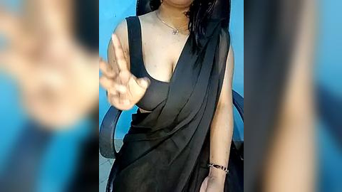 Media: A video of a woman with light brown skin and medium-length black hair, wearing a black sleeveless dress with a draped side, pointing at the camera, indoors with blue walls.