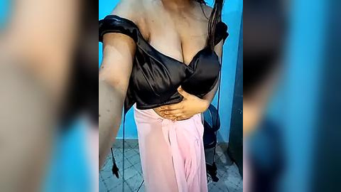 Media: Video of a light-skinned woman with large breasts, wearing a black, off-shoulder satin top and a pink skirt, standing in a blue-tiled room.