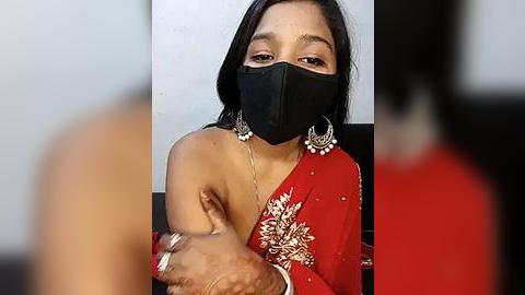 Media: Video of a South Asian woman with medium brown skin, wearing a red sari with gold embroidery, black face mask, and silver earrings. She is seated, with blurred figures in the background.