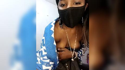 Media: Video of a Black woman with medium-dark skin, wearing a blue and white patterned robe, black face mask, and a silver necklace, revealing her large breast. Background is blurred.