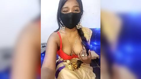 Media: A video of a South Asian woman with medium brown skin, wearing a red bra, black face mask, gold jewelry, and a beige sari, sitting on a blue couch, holding a large, erect penis.