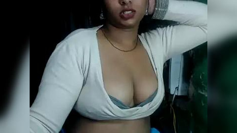 Media: Video of a Latina woman with medium skin tone, wearing a white cardigan and revealing a grey bra, her tongue out. Background is dimly lit with blurred objects.