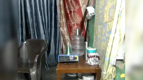 Media: Video of a small, cluttered room with a wooden desk holding a monitor, plastic bottles, and a framed picture. The background features colorful, patterned curtains and a white fan.